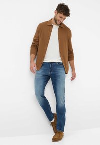 Cardigan marrone con bottoni sopra una t-shirt bianca, abbinato a jeans blu e stivali in suede marroni, con una vestibilità rilassata e una leggera texture sbiadita.