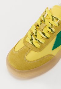 Yellow sneaker featuring a suede upper, fabric tongue, textured laces with black accents, and a transparent rubber sole. Green side logo detail.