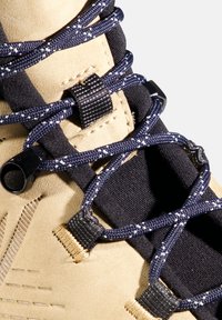 Tomaia in tessuto beige con base in tessuto nero, caratterizzata da lacci blu scuro intervallati da macchie bianche e dettagli hardware neri.