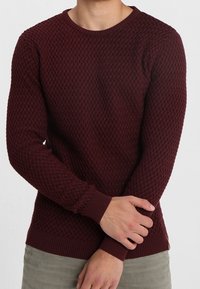 Burgundy texturerad stickad tröja med en rundad halsringning och ribbade mudd, visad på en person med armarna korsade.