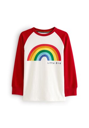 Enfant assis en tailleur portant un t-shirt blanc avec des manches rouges et un graphique arc-en-ciel multicolore qui dit "Little Bird."