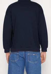 Marinblå bomulls sweatshirt med långa ärmar och ribbade ärmslut, med en avslappnad passform och sömnadsdetaljer. Bärs med blå denimshorts.
