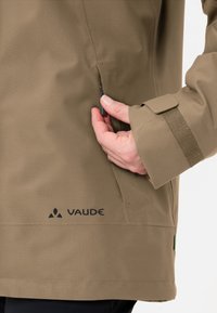 Giacca verde oliva con una texture di tessuto liscia, tasca laterale con zip, polsini regolabili e logo "Vaude" nero sulla parte bassa davanti.