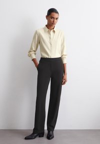 Cămașă crem cu guler, cu mâneci lungi, asortată cu pantaloni negri cu talie înaltă. Modelul poartă cizme negre până la gleznă pe un fundal neutru.