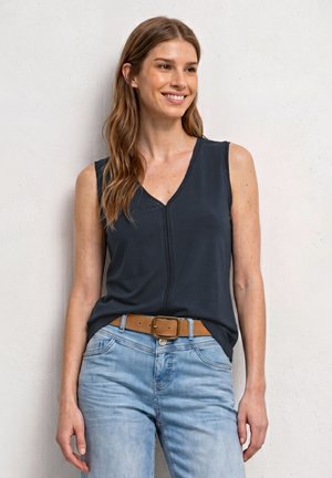 Femme souriante aux longs cheveux portant un haut sombre sans manches, un jean bleu clair et une ceinture marron, debout contre un mur clair.