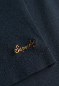Gros plan sur un tissu marine avec un logo "Superdry" brodé en brun près du bord cousu.