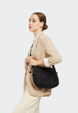 Black patterned shoulder bag with a smooth texture, featuring dual zipper pockets and an adjustable strap, carried against a beige outfit.