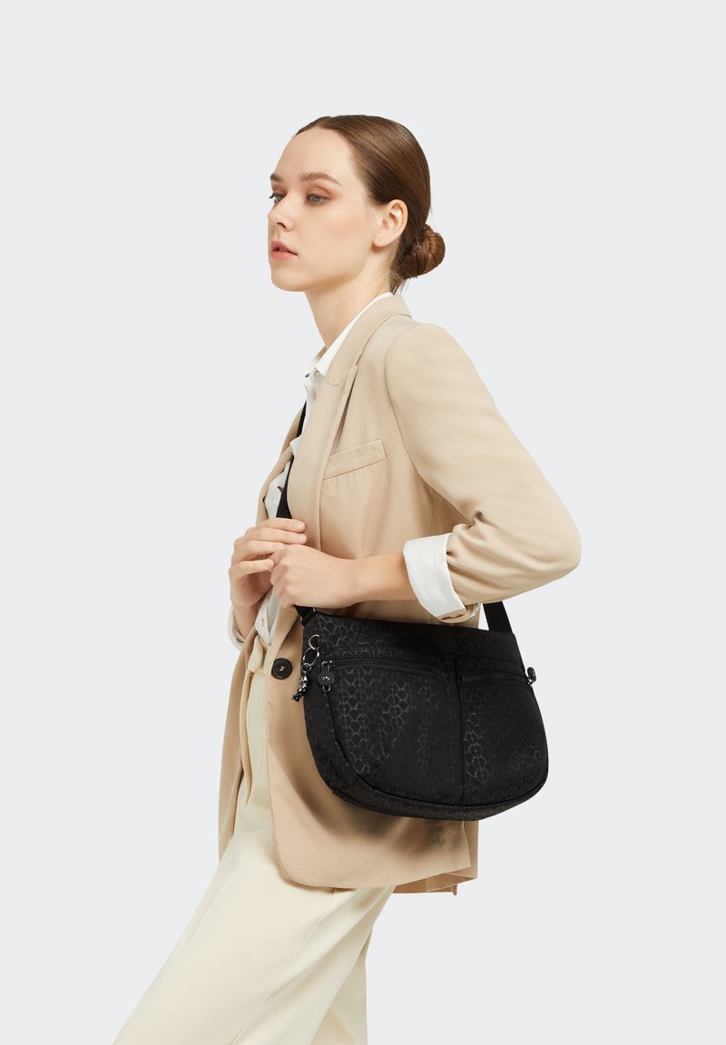 Black patterned shoulder bag with a smooth texture, featuring dual zipper pockets and an adjustable strap, carried against a beige outfit.