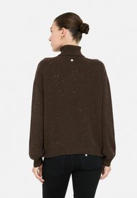 Vrouw met haar in een knot, gekleed in een bruin gemêleerd coltrui en zwarte jeans, van achteren weergegeven tegen een plain witte achtergrond.