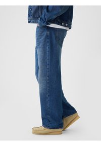 Jean en denim bleu à coupe décontractée, avec un lavage clair, un léger effet délavé et des ourlets cousus, assorti à des chaussures style mocassin beige.