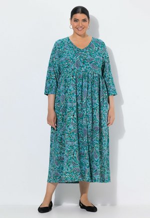 Kobieta ubrana w turkusową sukienkę midi w paisley z rękawami do łokcia i czarne baleriny, stojąca na białym tle, uśmiechająca się.