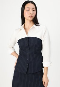 Cămașă albă cu guler superior, cu un corset în dungi bleumarin, având nasturi și o croială structurată. Designul include un detaliu cu helancă.