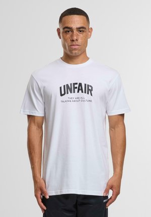 Hvid bomulds t-shirt med fed sort tekst "UNFAIR" og sætningen "DE TALER ALLE OM KULTUR" nedenfor. Kortærmet og med rund hals.