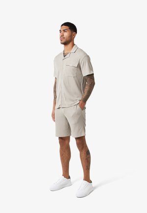 Homme debout, mains dans les poches, portant une chemise beige à manches courtes boutonnée et un short assorti, avec des baskets blanches sur fond blanc.