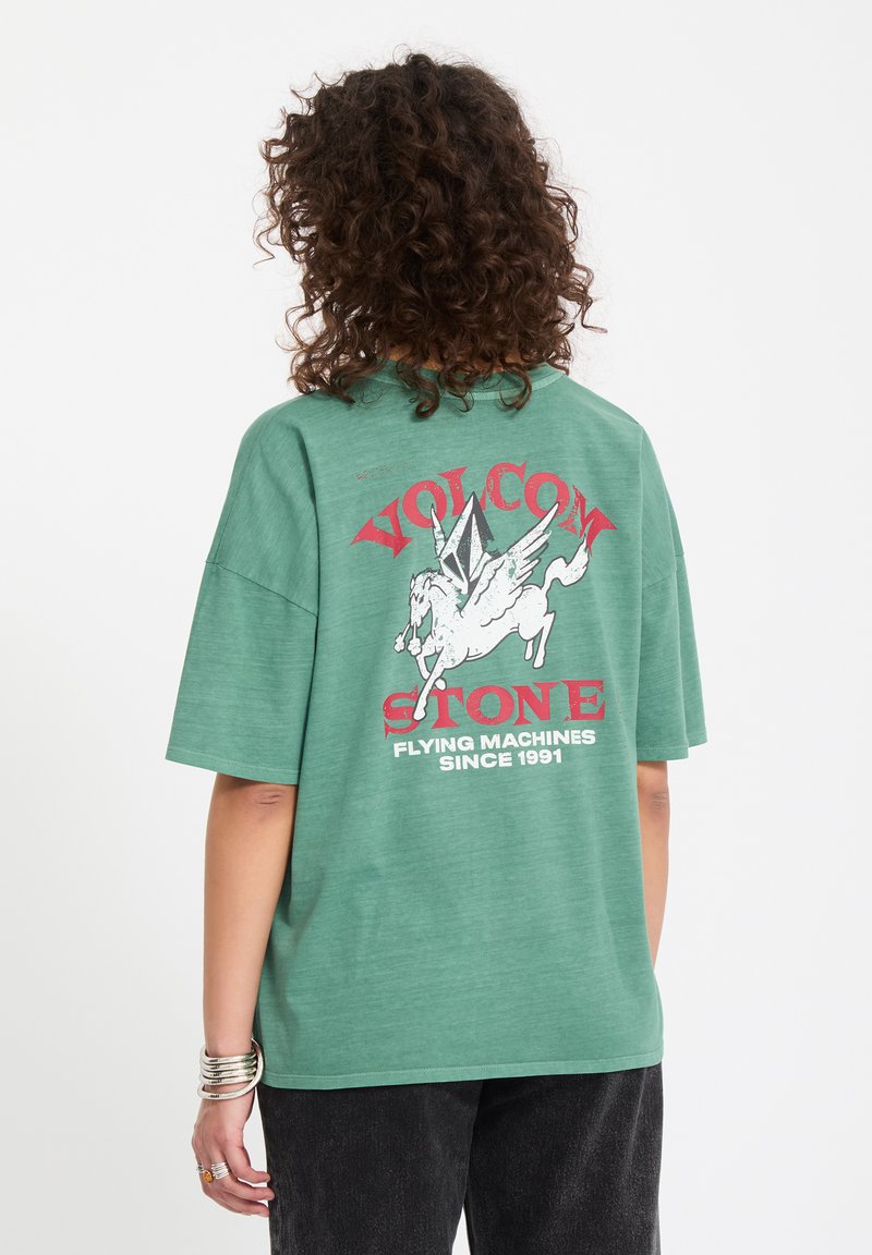 Personne aux cheveux bouclés portant un t-shirt vert avec le logo du cheval ailé Volcom Stone rouge et blanc ainsi qu'un texte au dos.