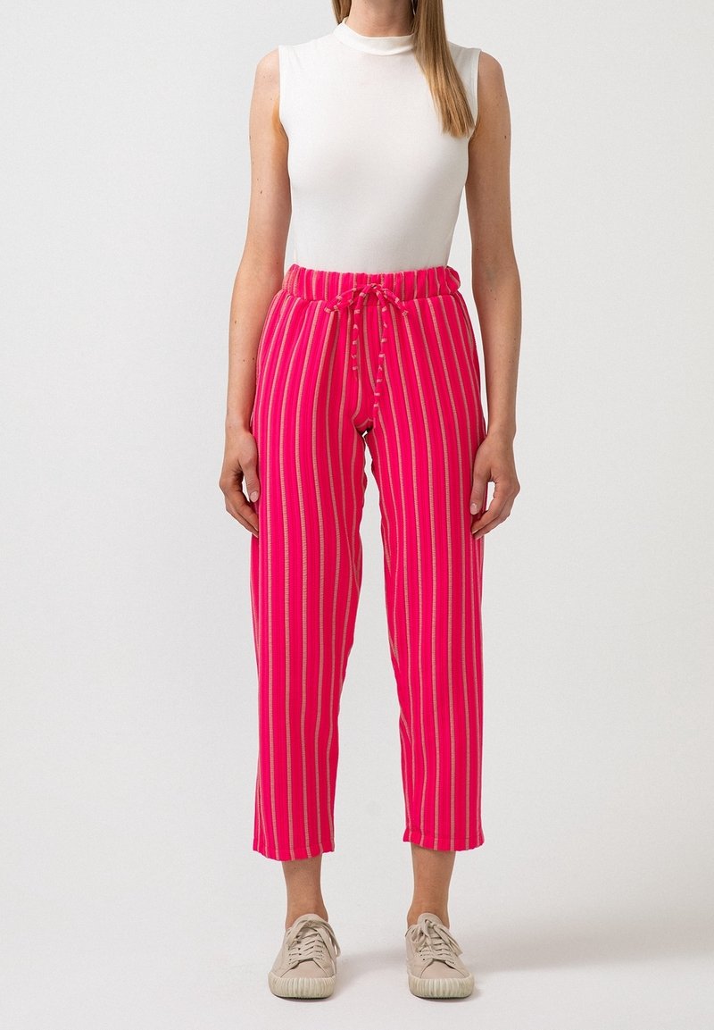 Femme portant un pantalon rayé rose vif avec une taille à cordon, un débardeur blanc et des baskets beiges, debout devant un fond uni.