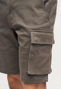 Καφέ cargo σορτς με μεγάλη τσέπη με καπακι στην πλευρά, φορεμένα από ένα άτομο, με λεπτομερή ραφές και υφή υφάσματος.