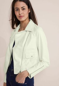 Veste en simili cuir vert clair avec col rabattu, fermeture éclair, deux poches avant et poignets zippés. Portée par-dessus un haut sombre.