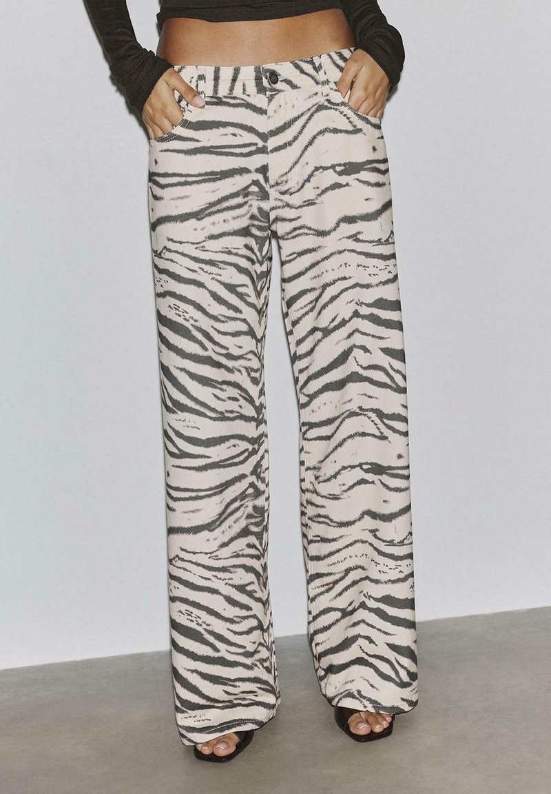 Femme portant un pantalon taille haute à jambes larges avec un motif zèbre gris et blanc, debout les mains dans les poches.