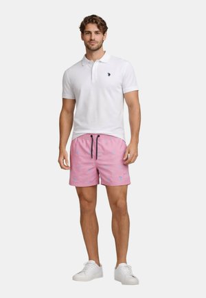 Mann trägt weißes Poloshirt, pinke Badeshorts mit blauen Mustern und schwarzen Kordeln sowie weiße Sneakers, steht vor neutralem Hintergrund.