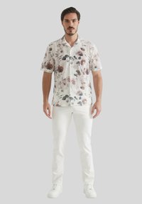 Chemise à manches courtes à fleurs sur un fond blanc, avec des motifs rouge et vert atténués, assortie à un pantalon blanc et des baskets.
