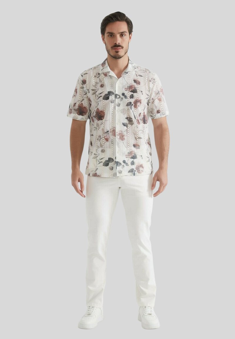 Chemise à manches courtes à fleurs sur un fond blanc, avec des motifs rouge et vert atténués, assortie à un pantalon blanc et des baskets.