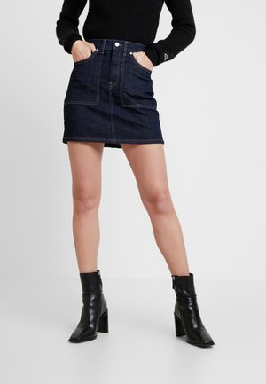 Woman wearing a dark denim mini skirt, black sweater, and black leather ankle boots with block heels, standing with hands in skirt pockets.