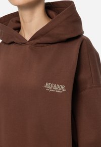 Sudadera marrón con una capucha grande, textura suave y un pequeño logo bordado en color claro en el pecho. Diseño simple y casual.