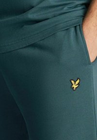 Pantaloni jogger aderenti color verde petrolio, con piccolo logo di aquila gialla ricamata sul lato e tessuto dalla texture liscia.