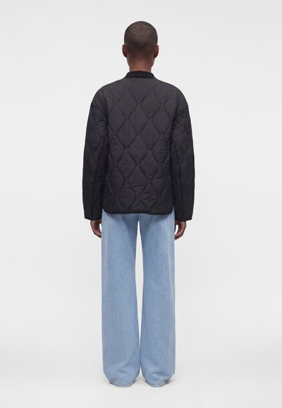 Personne de dos, portant une veste matelassée noire et un jean bleu clair à jambes larges, debout contre un fond blanc uni.