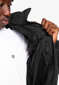 Sort lynlåsjakke med en glat overflade og høj krave, båret over en hvid t-shirt med et lille logo. Inderlommen er synlig.