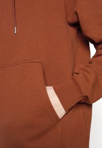 Sweat-shirt couleur rouille avec poche kangourou, tissu texturé et capuche avec cordon de serrage. Comprend des poignets et un ourlet côtelés.