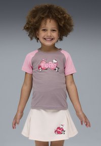 Uśmiechnięte dziecko z kręconymi włosami, ubrana w różową i szaro-czarną koszulkę Puma z motywem Hello Kitty oraz białą spódniczkę z pasującym wzorem Hello Kitty.