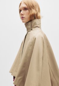 Giacca a mantellina beige con colletto alto, design strutturato e texture liscia. Presenta una zip frontale e una silhouette fluida.