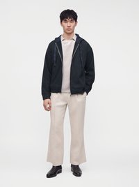 Jeune homme debout portant un sweat à capuche noir zippé, un polo beige clair, un pantalon large beige et des chaussures habillées noires sur fond blanc.