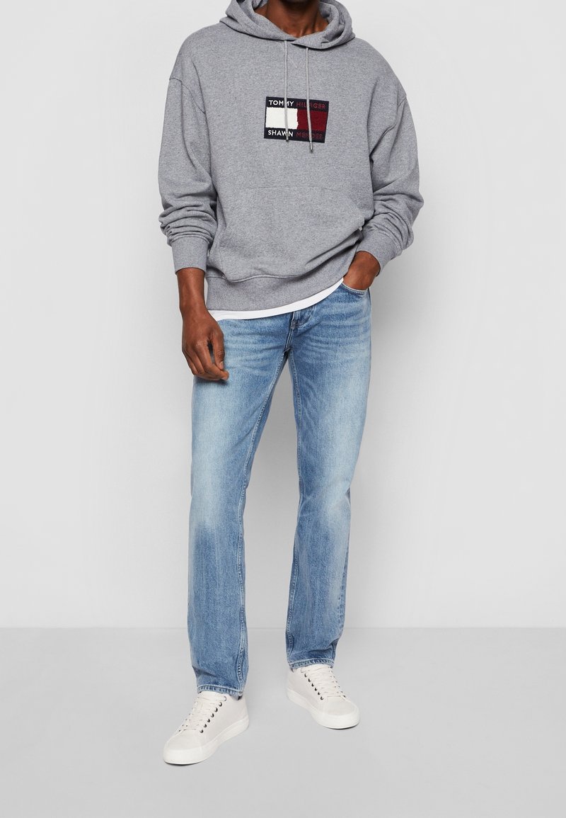 Sudadera gris con parche de logo, combinada con jeans de mezclilla azul claro y zapatillas blancas. Presenta capucha con cordón y un corte relajado.