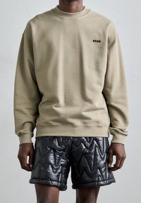 Felpa khaki con polsini e scollo a costine, con un logo nero sul petto. Abbinata a pantaloni shorts neri lucidi e trapuntati.
