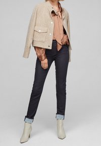 Chaqueta de tweed beige con botones plateados y dos bolsillos frontales, combinada con una blusa cobriza y jeans oscuros, terminada con botas de tacón de un beige claro.