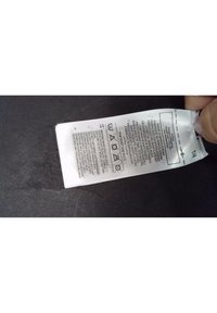 Biała metka pielęgnacyjna z instrukcjami prania, tekst w wielu językach i symbole pielęgnacyjne, przymocowana u góry i widoczna na ręce na dole.