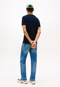 Man met een zwart T-shirt, blauwe spijkerbroek, witte sneakers en een groene pet staat met zijn rug naar ons toe en heeft zijn handen achter zijn rug in elkaar gevouwen.