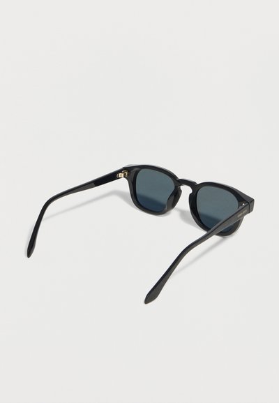 Γυαλιά ηλίου με μαύρο πλαίσιο και στρογγυλούς σκοτεινούς φακούς, ανοιχτά πάνω σε μια απλή ανοιχτόχρωμη επιφάνεια, με τα δύο μπράτσα επεκταμένα.