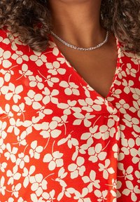 Tela roja con patrón floral blanco, con un diseño de escote en V. Se puede ver un collar de cadena plateada contra el escote.
