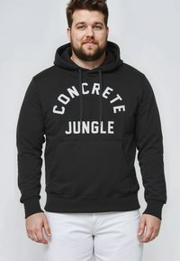 Sudadera negra con texto blanco "CONCRETE JUNGLE" en un arco. Presenta una capucha con cordón y un bolsillo frontal tipo canguro, fabricada con tejido suave.