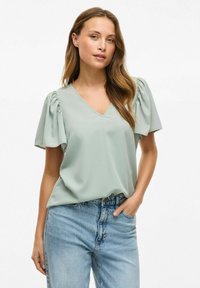 Svijetlozelena majica kratkih rukava s V-izrezom i napuhanim rukavima. Izrađena od mekog materijala s glatkom teksturom. Uparena s trapericama visokog struka.