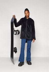Flauschige schwarze Zip-Jacke mit Kapuze, blaue Schneehose und schwarze Stiefel. Hält ein Snowboard mit Bindungen. Schlichtes und funktionales Design.