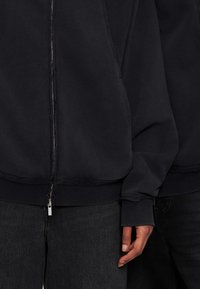 Κοντινό πλάνο δύο ατόμων που φορούν μαύρες φανέλες με φερμουάρ και μαύρα τζιν, με το χέρι του ενός να φαίνεται δίπλα στο σώμα του.