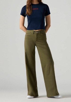 Vrouw draagt een cropped marineblauw T-shirt met rode tekst, gecombineerd met olijfgroene, wijde broek met hoge taille en witte schoenen.