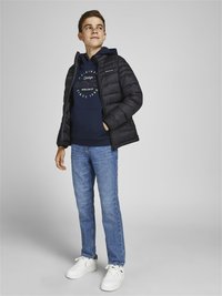 Giubbotto puffo nero con zip frontale, felpa blu navy con stampa grafica, jeans azzurri e sneakers bianche. Tessuto morbido, vestibilità casual.