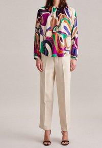 Mångfärgad blus med abstrakta mönster i lila, orange och grönt, kombinerad med krämfärgade skräddarsydda byxor. Svarta öppna klackskor.