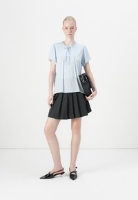 Blusa azzurro chiaro a maniche corte con colletto arricciato, abbinata a una gonna plissettata nera e tacchi slingback neri. Tiene una borsa nera.